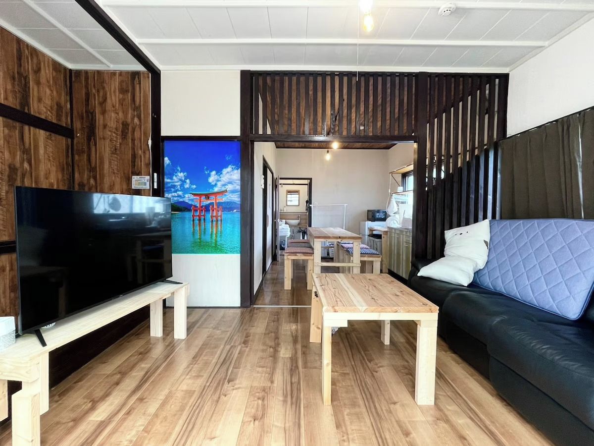 Ein modernes Wohnzimmer mit Flachbildfernseher, Holzboden, einer Couch mit einer gesteppten Rückenlehne und einem Essbereich mit Holztischen und -stühlen.