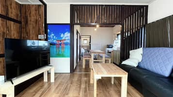 Ein modernes Wohnzimmer mit Flachbildfernseher, Holzboden, einer Couch mit einer gesteppten Rückenlehne und einem Essbereich mit Holztischen und -stühlen.