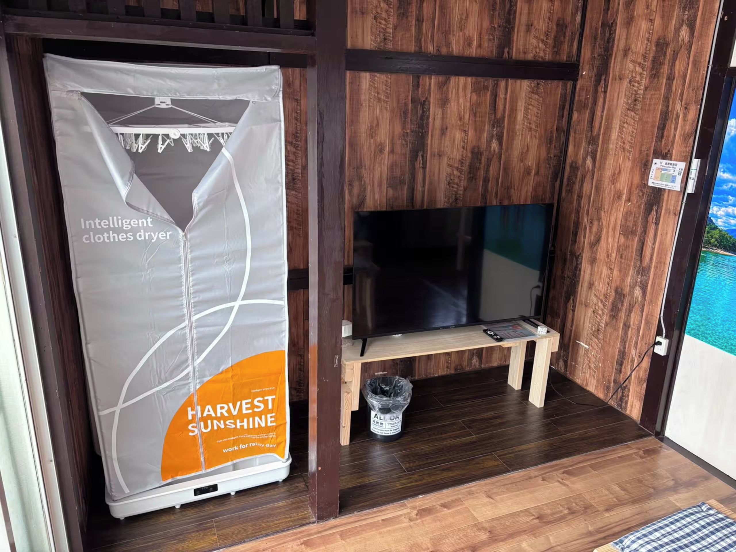 Ein Zimmer mit Holzboden, einem Fernseher auf einem Holzständer und einem Wäschetrockner mit den Aufschriften 'Intelligent clothes dryer' und 'Harvest Sunshine'.