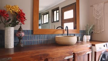 A bathroom with a wooden vanity, a sink with a vessel basin, a mirror, a washing machine, and a macrame wall hanging.