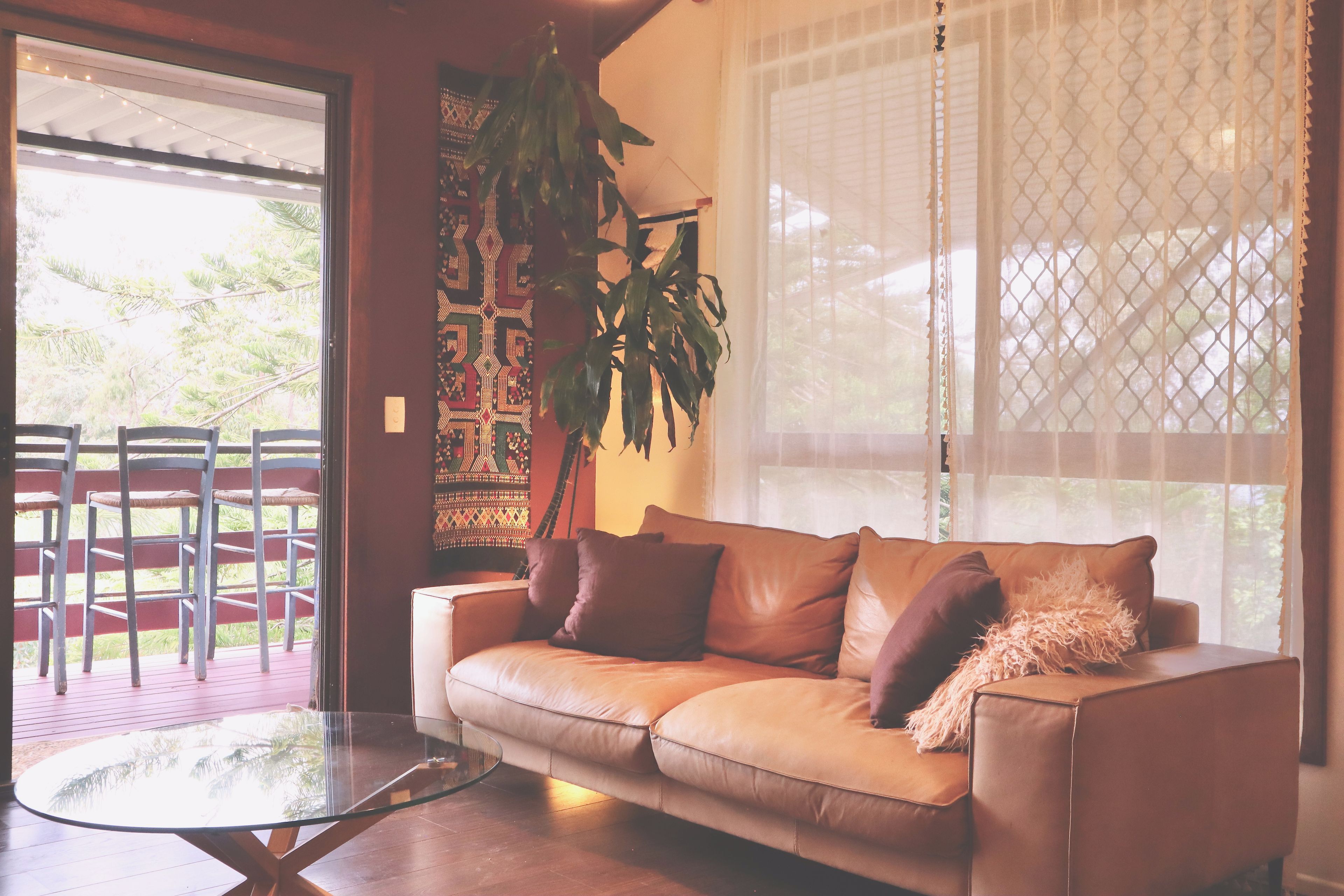 A room with a glass-top table, a brown leather sofa with cushions, a hanging plant, and a view through a window with a white curtain.