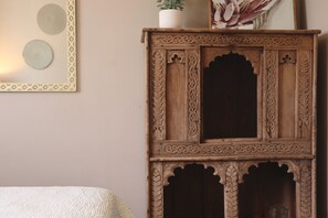 A wooden cabinet with intricate carvings, a framed picture above it, and a bed with a textured blanket.