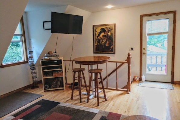 Una habitación con suelo de madera, una mesa redonda con taburetes, un televisor montado en la pared, una estantería y una puerta con vistas.