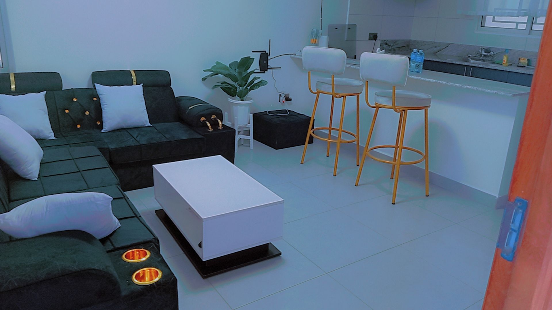 A modern living room with a sofa, ottoman, and a coffee table. There are two bar stools and a small kitchen area with a sink and countertop.