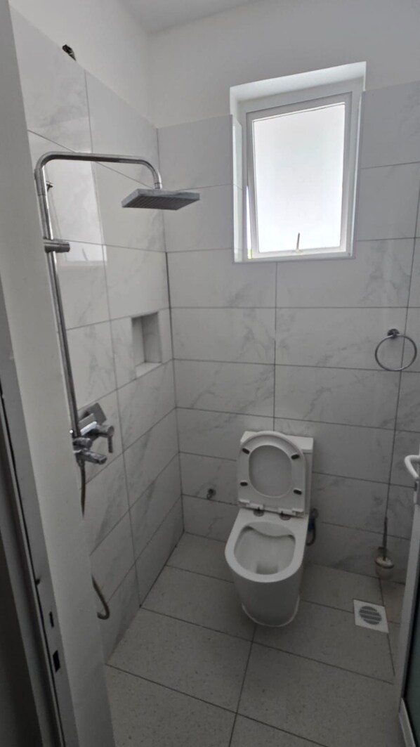 A small, tiled bathroom with a toilet, a shower, and a window. -  Dolphin Court Apartment – Stylish 2-Bedroom Stay Near Diani Beach (Diani Beach)