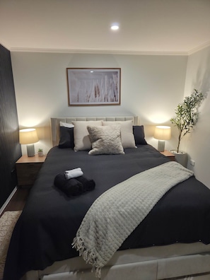 Ein ordentlich eingerichtetes Schlafzimmer mit einem großen Bett, zwei Nachttischlampen, einem Wandbild und einem Topf mit einer Pflanze.