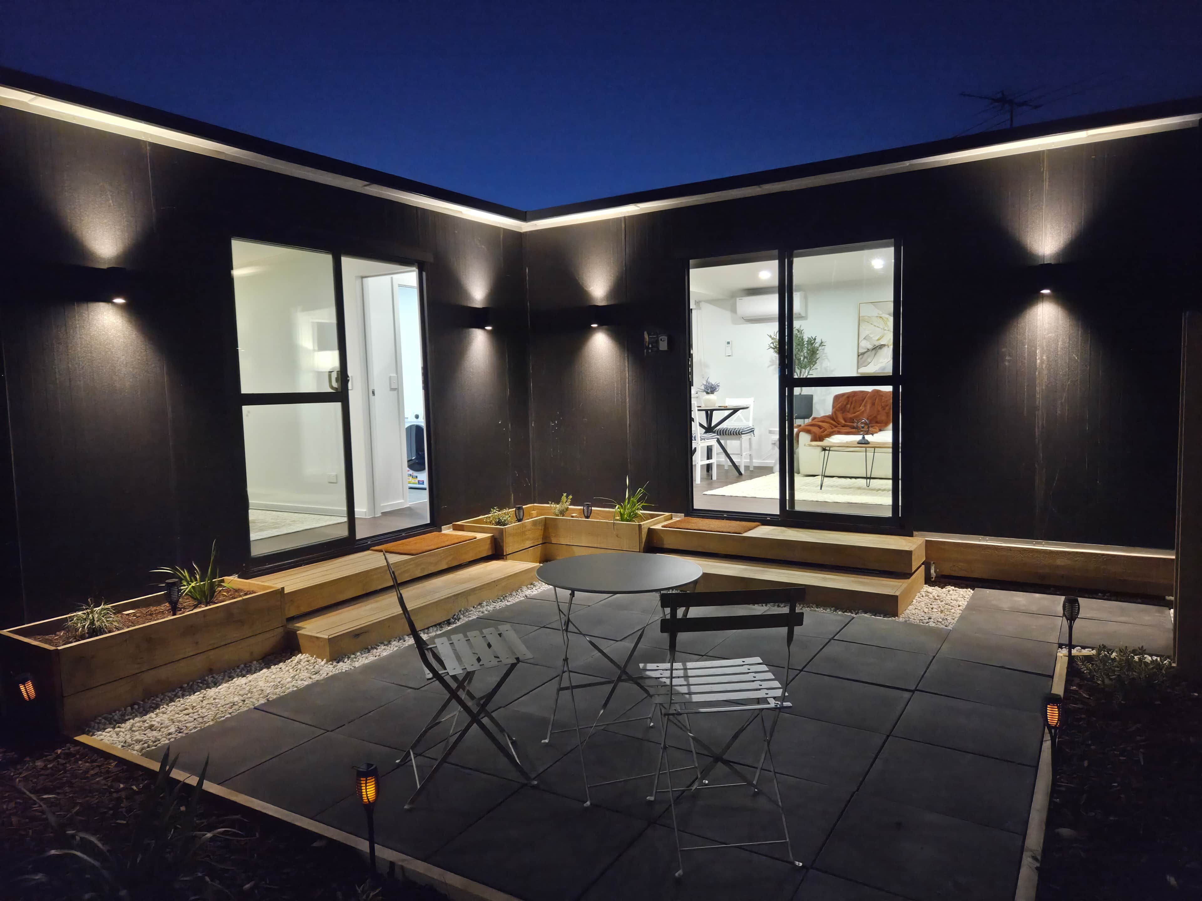 Zona relax esterna moderna con tavolo e sedie, circondata da pareti scure e ampie finestre.
