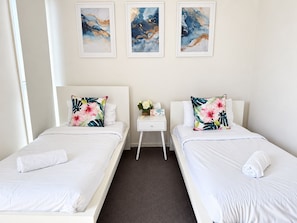 A hotel room with two beds, white bedding, floral pillows, a small bedside table with a vase of flowers, and abstract art on the wall. - Waterfront 2BR Townhouse | Balcony & parking (Carrara)