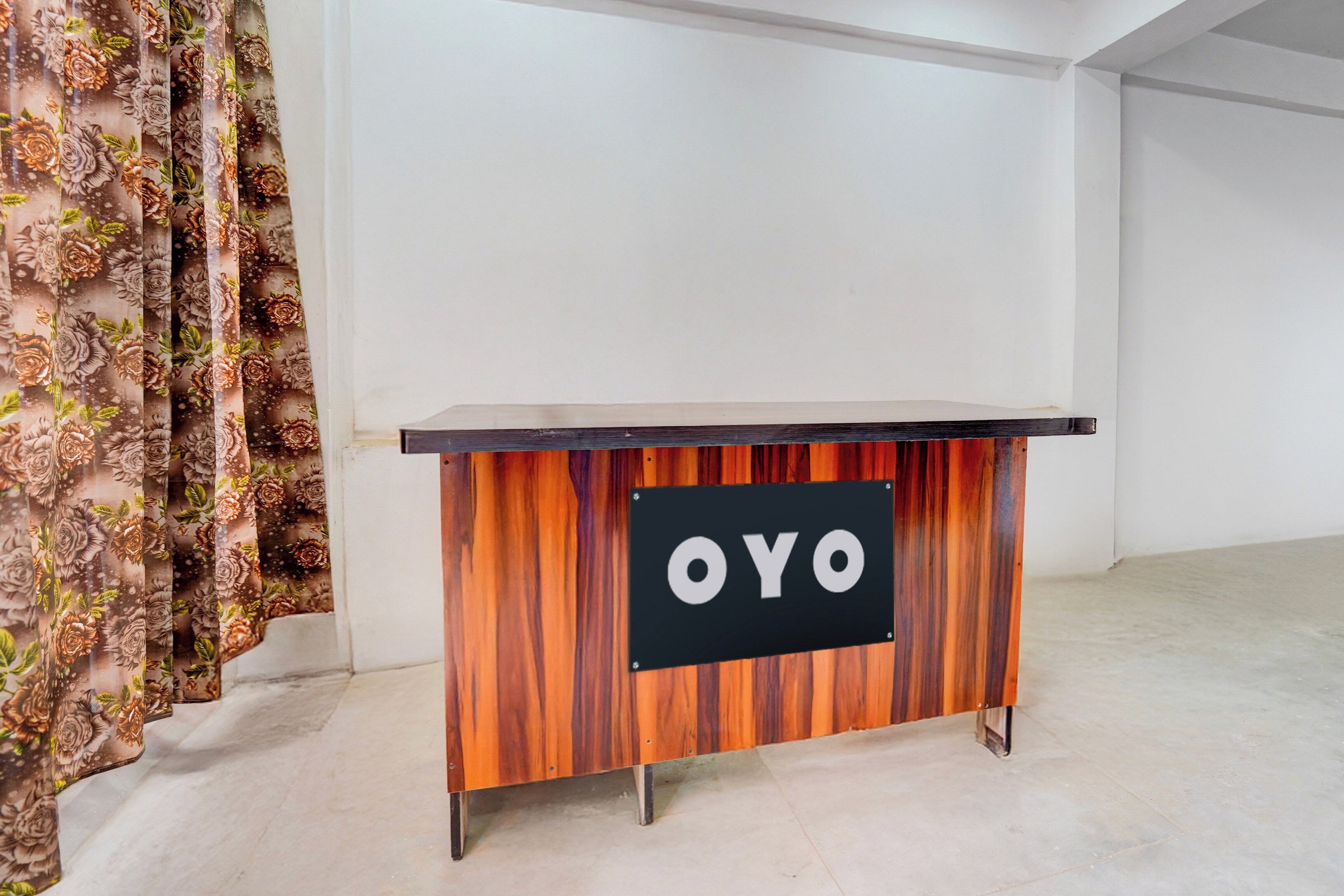 Un bureau en bois avec un panneau noir sur lequel est inscrit en lettres blanches le mot « OYO », situé dans une pièce dont le mur est d’un blanc uni et qui est agrémentée d’un rideau à motifs floraux.