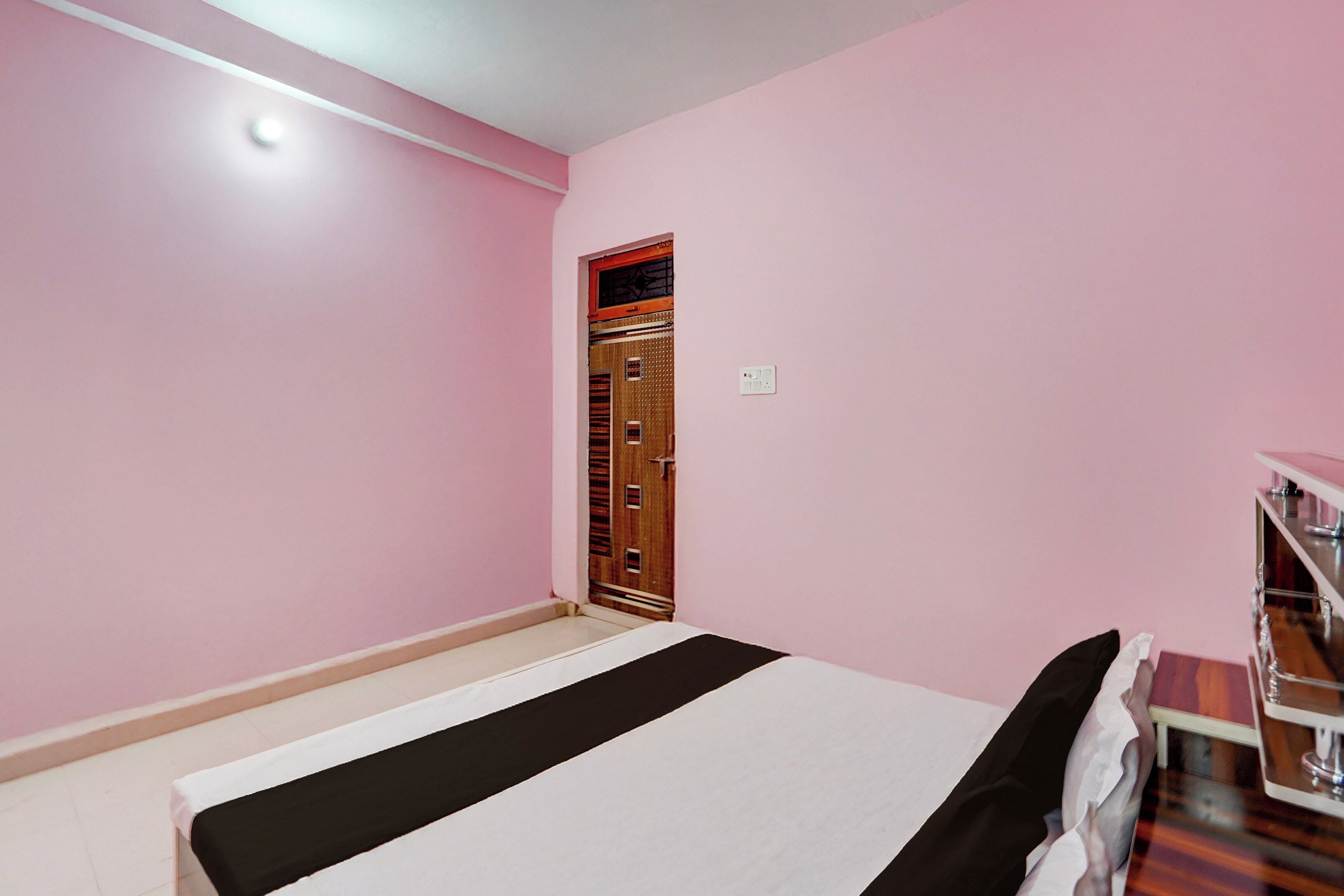 Una habitación con paredes rosas, una cama con colcha blanco y negro, una puerta de madera y un enchufe blanco en la pared.