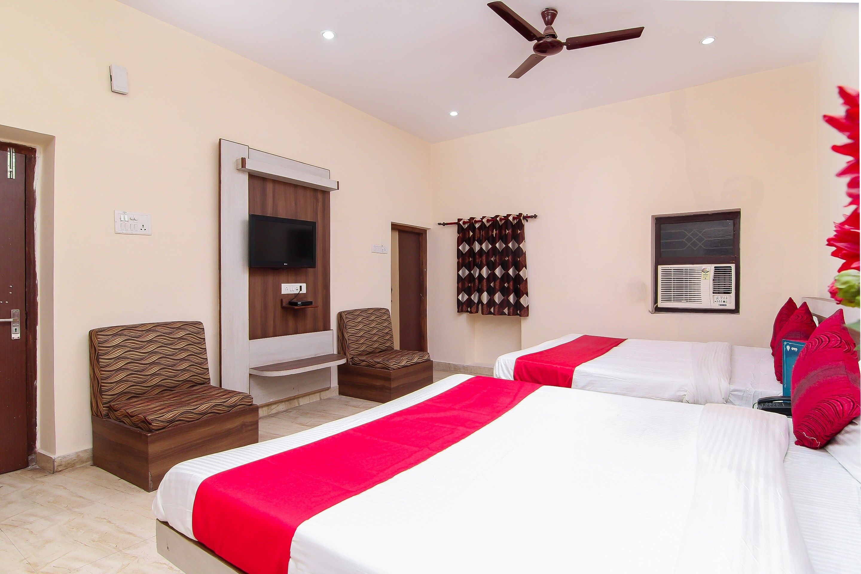 Habitación de hotel con dos camas, ventilador de techo, televisor y ventana con aire acondicionado.