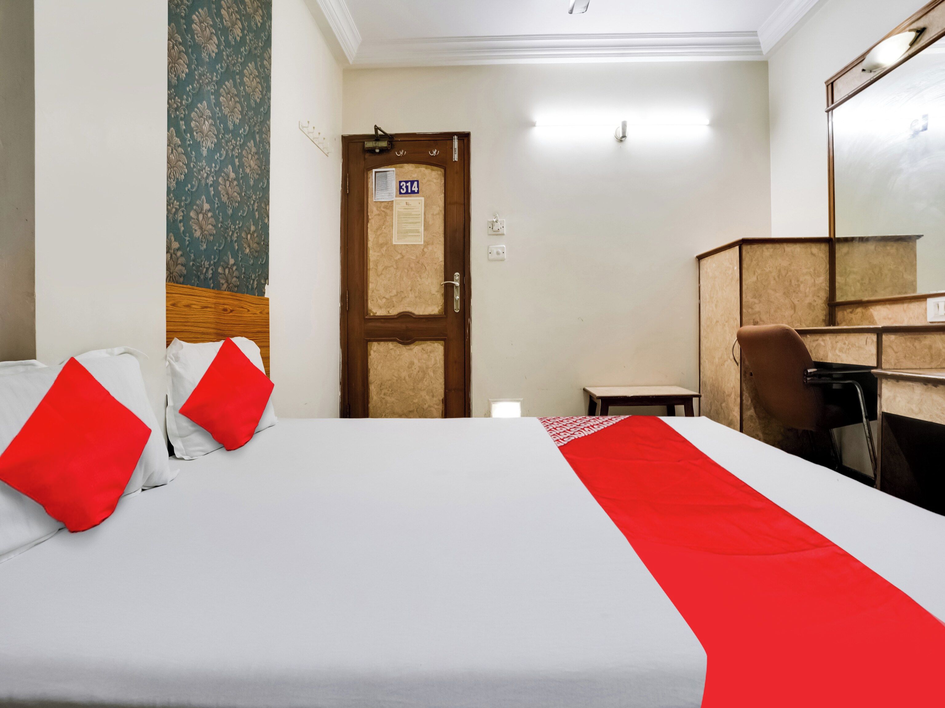 A hotel room with a bed, a desk, a chair, and a door with a number sign.