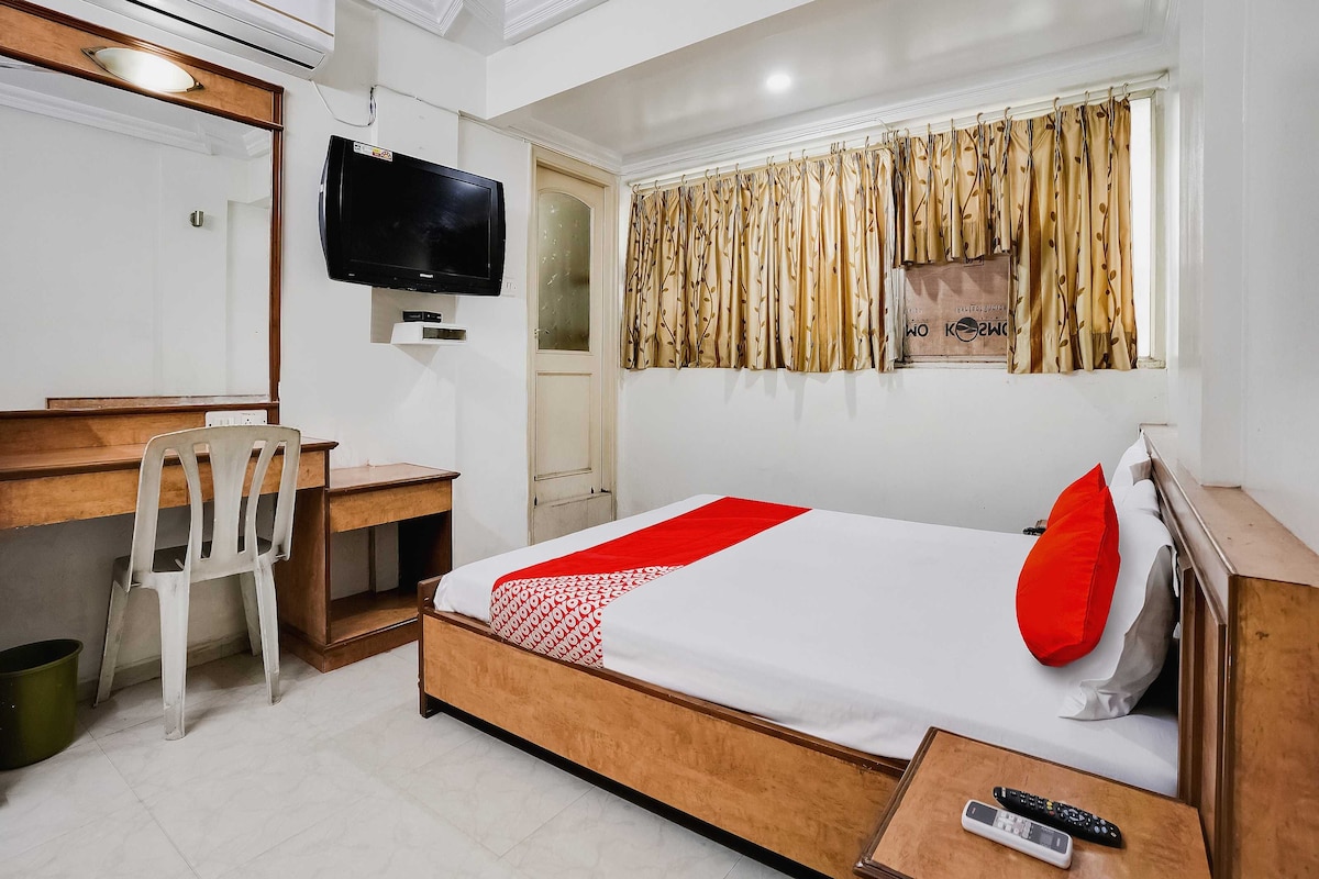 Habitación de hotel con cama, escritorio, silla, televisor y ventana con cortinas.