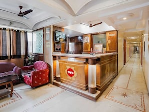 A reception area with a reception desk, a red sofa, a wooden chair, and a ceiling fan. - Hotel O Raj Palace (Ahmedabad)