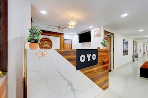 A hotel reception desk with a modern design, a flat-screen TV, and a sign indicating the location of the pantry. - Hotel O by OYO Shaligram (Ahmedabad)