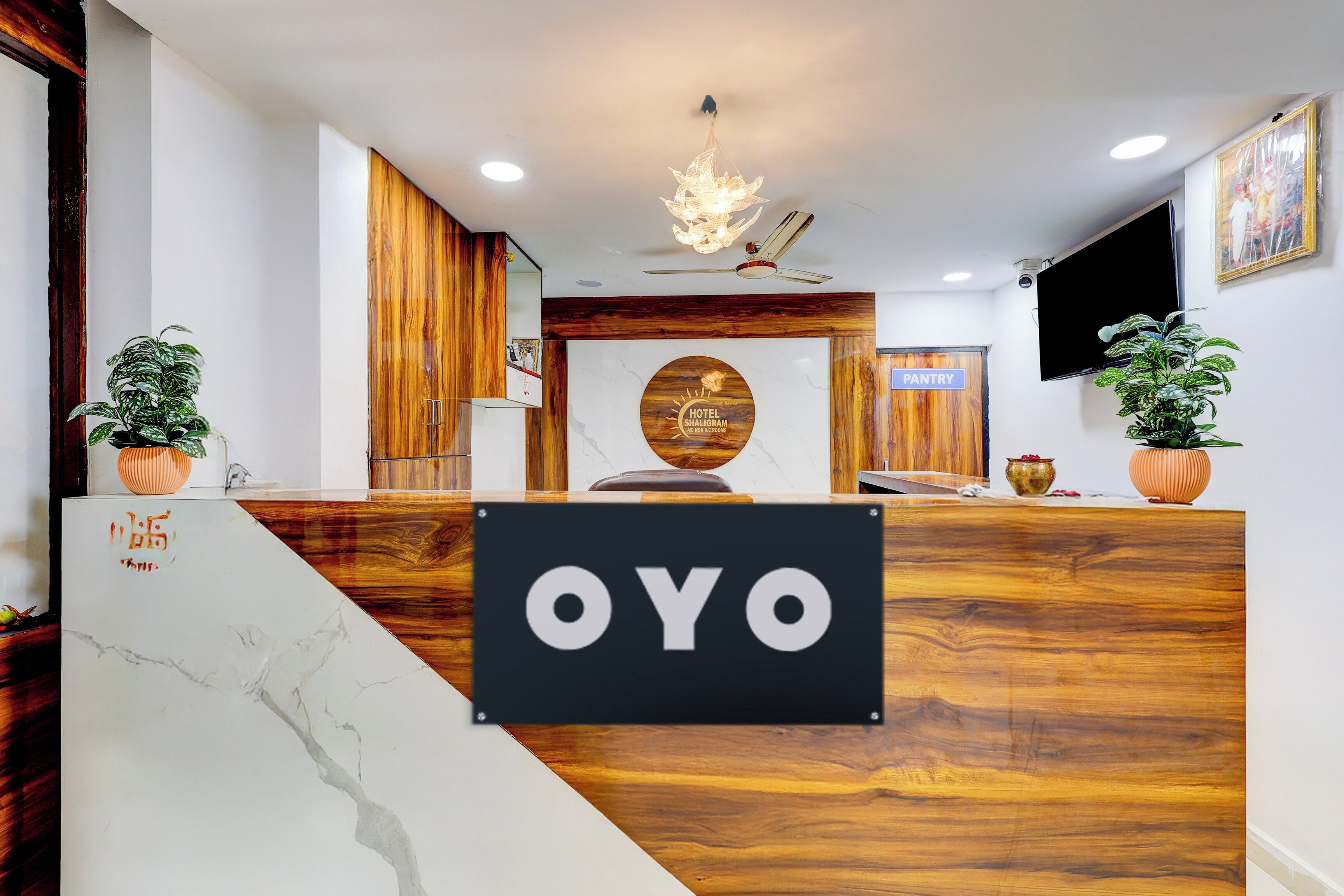 A reception desk with a sign reading 'OYO' in a hotel lobby with wooden accents, a chandelier, and a television on the wall.