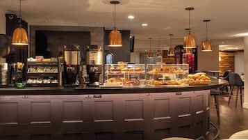 A modern bar area with a counter displaying various food items, pendant lighting, and a seating area with round tables.