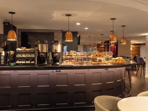 Eine moderne Bar mit einer Theke, auf der verschiedene Speisen präsentiert werden, Pendelleuchten und einer Sitzgelegenheit mit runden Tischen.