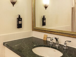 Ein Badezimmer mit Marmorwaschbecken, zwei Wasserhähnen, einem Spiegel mit goldener Rahmung und zwei Seifenspendern an der Wand.
