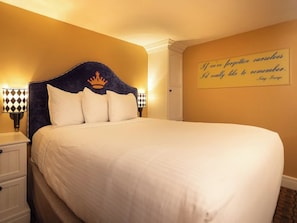 Ein ordentlich bezogenes Bett mit einem an der Bettstatt befestigten Kopfteil mit Kronenmuster, eine Nachttischlampe mit kariertem Lampenschirm und ein motivierendes Zitat an der Wand.