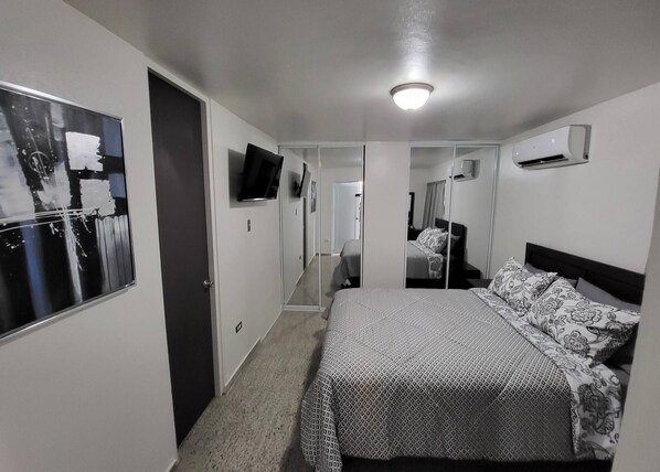 A hotel room with a bed, a TV mounted on the wall, a door, and an air conditioning unit.