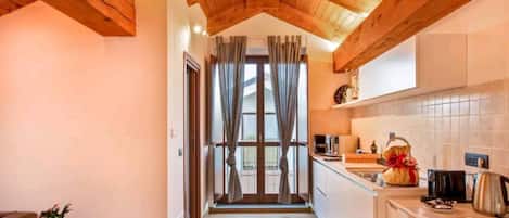A narrow hallway leading to a kitchen with wooden beams, a small table with a plant, and a window with curtains.