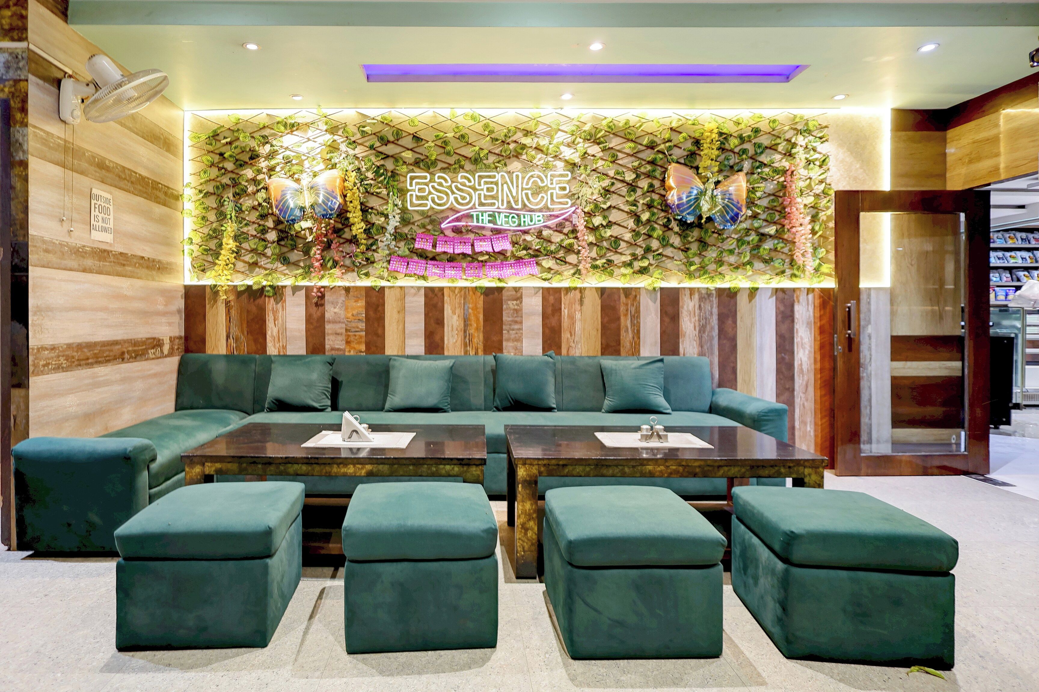 A modern lounge area with a green sofa, matching ottomans, and a wall-mounted sign reading 'ESSENCE THE VEG HUB'.