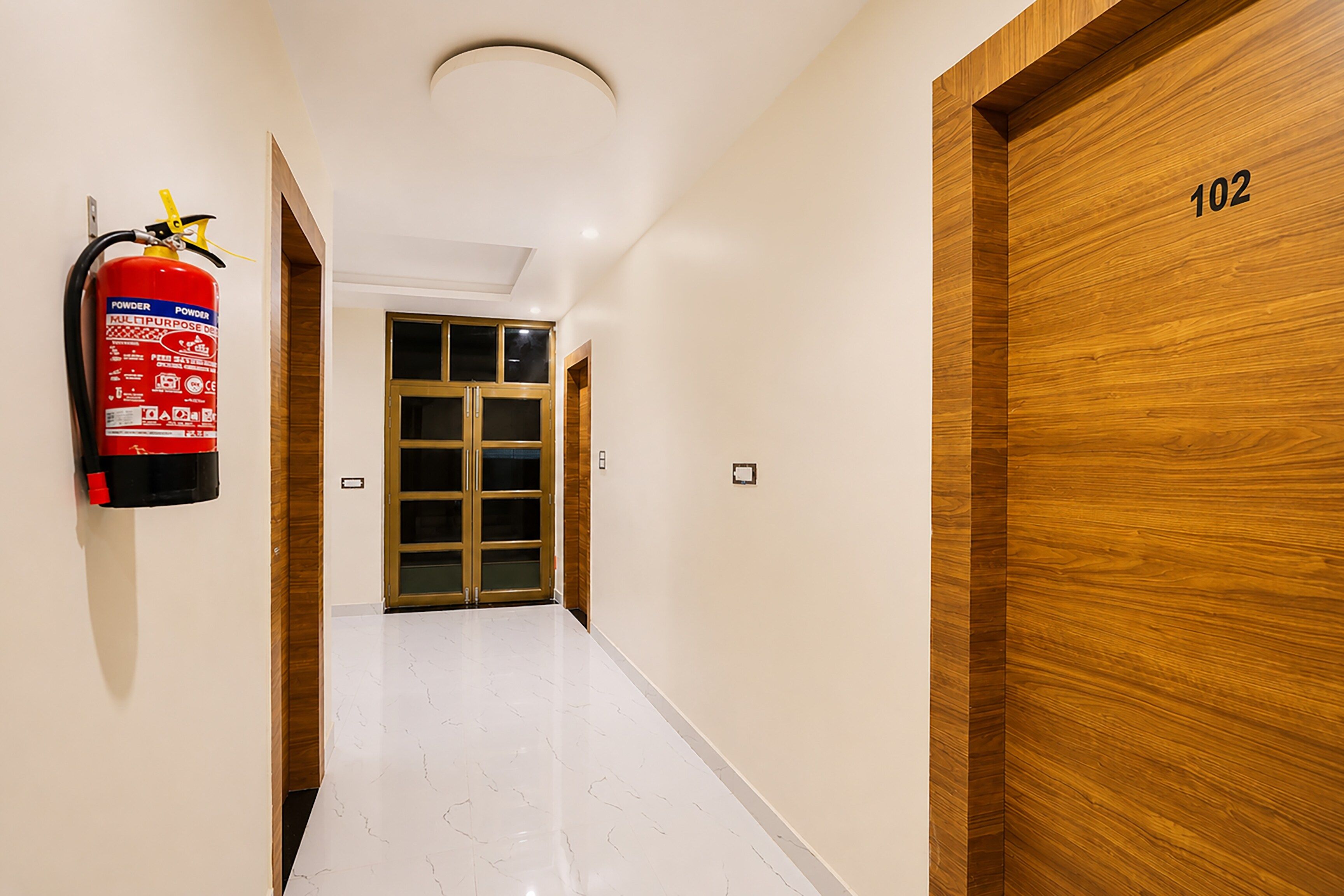 A hallway with a fire extinguisher mounted on the wall, a wooden door with the number 102, and a double door at the end.