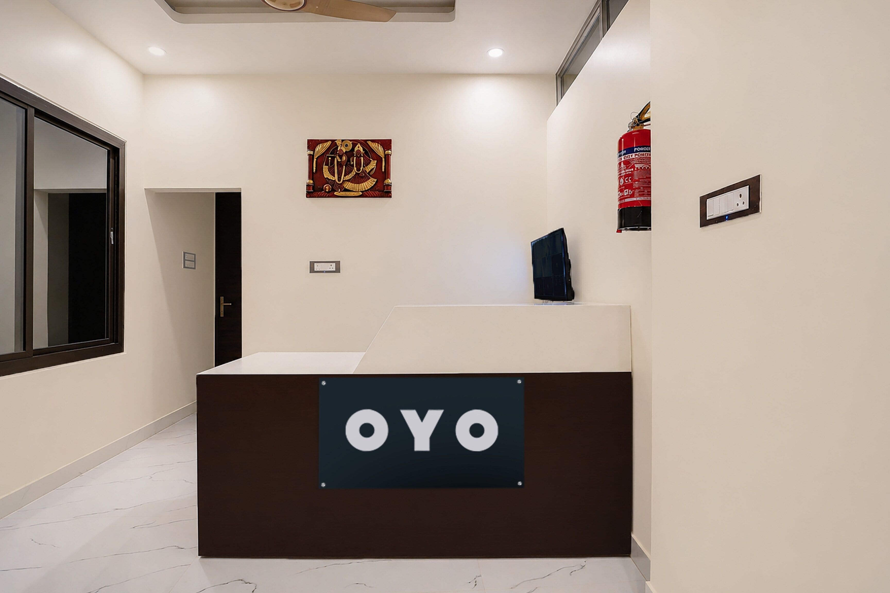 A reception desk with a sign that reads 'OYO' in a modern interior with a ceiling fan, a fire extinguisher, and a framed picture on the wall.