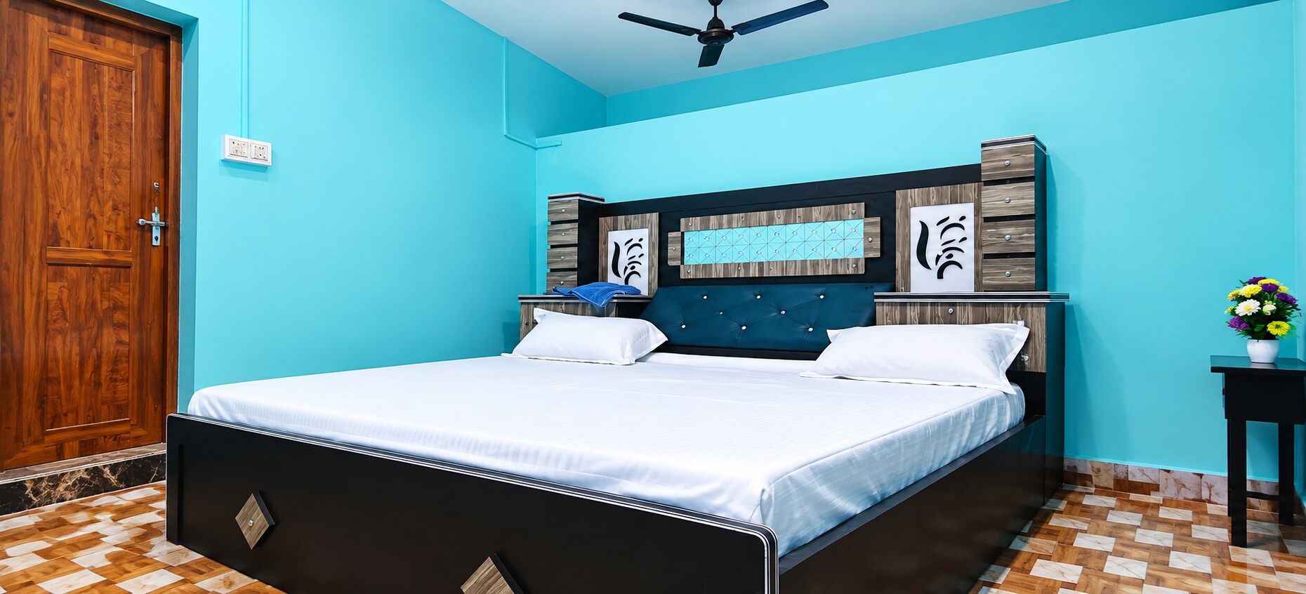 A bedroom with a bed, a wooden door, a ceiling fan, a nightstand, and a vase with flowers.