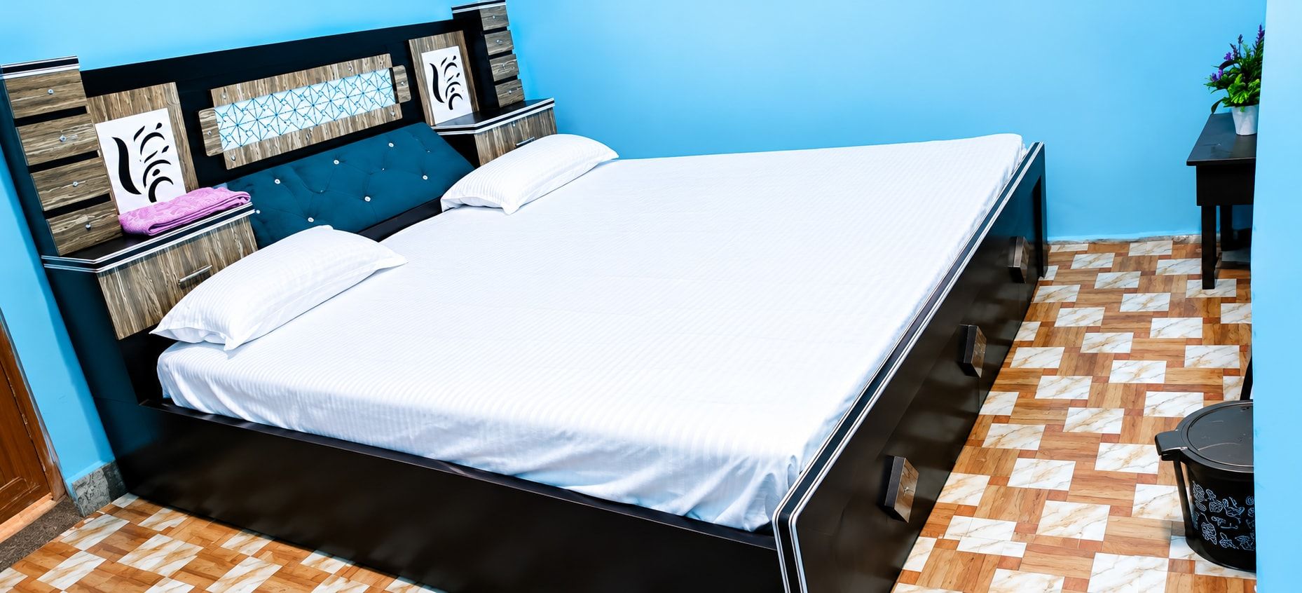 A bed with a white mattress and a dark headboard, a wooden nightstand with a pink towel, and a potted plant on a blue wall.