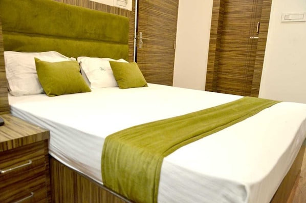 Una cama bien tendida con cabecera verde y ropa de cama blanca, una mesita de noche con cajones y un armario de madera con tirador.