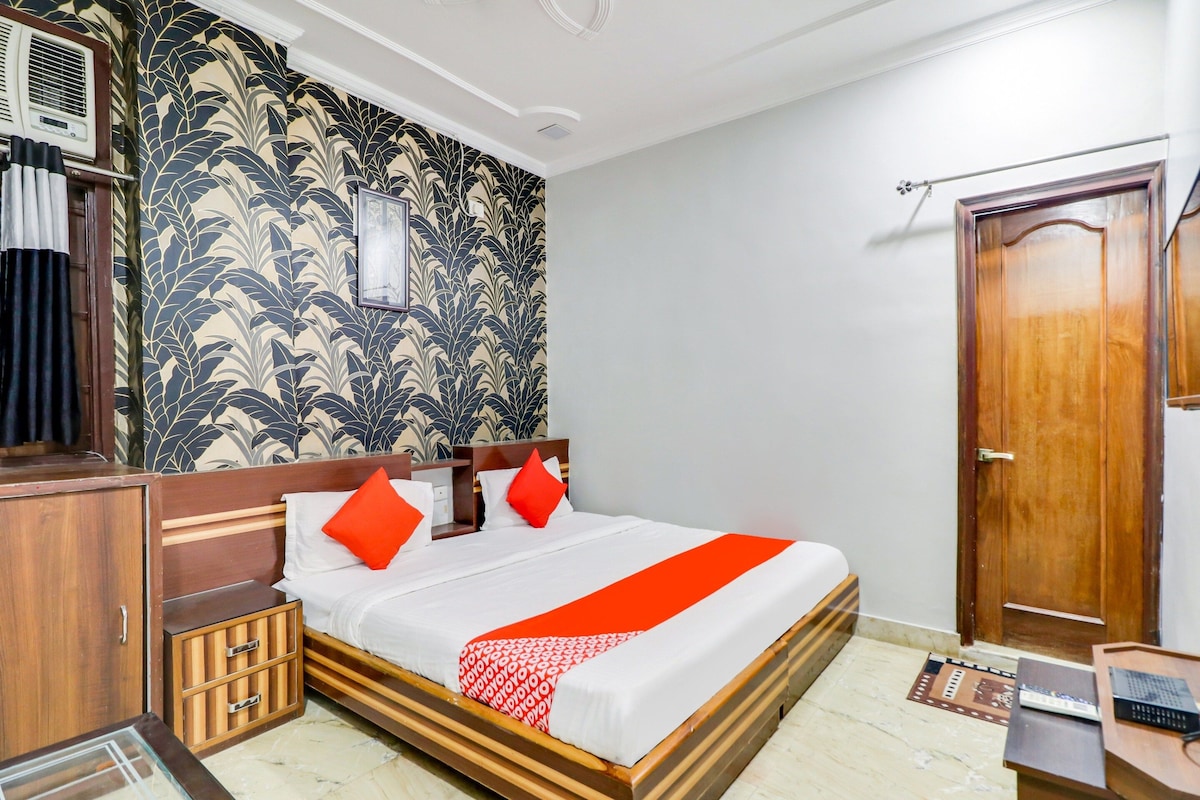 A hotel room with a bed, wooden headboard, a wardrobe, a mirror, a door, and a small table with a remote control.