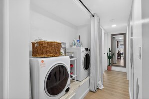 A laundry room with two washing machines, a wicker basket on the dryer, and a potted plant. - The Velvet Cactus, Private Pool, Fire Pit, BBQ, & King Bed (Indio)