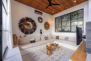 A modern living room with a wooden ceiling, a large window with a view, a sofa with patterned cushions, and decorative wall mirrors. - 2BR Private Pool Villa with Rice Field Views & Sunset Balconies (Badung)