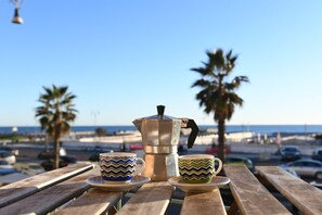 A wooden table with a coffee maker and two patterned cups, overlooking a beach with palm trees and a clear sky. - New Listing (Lido di Ostia)
