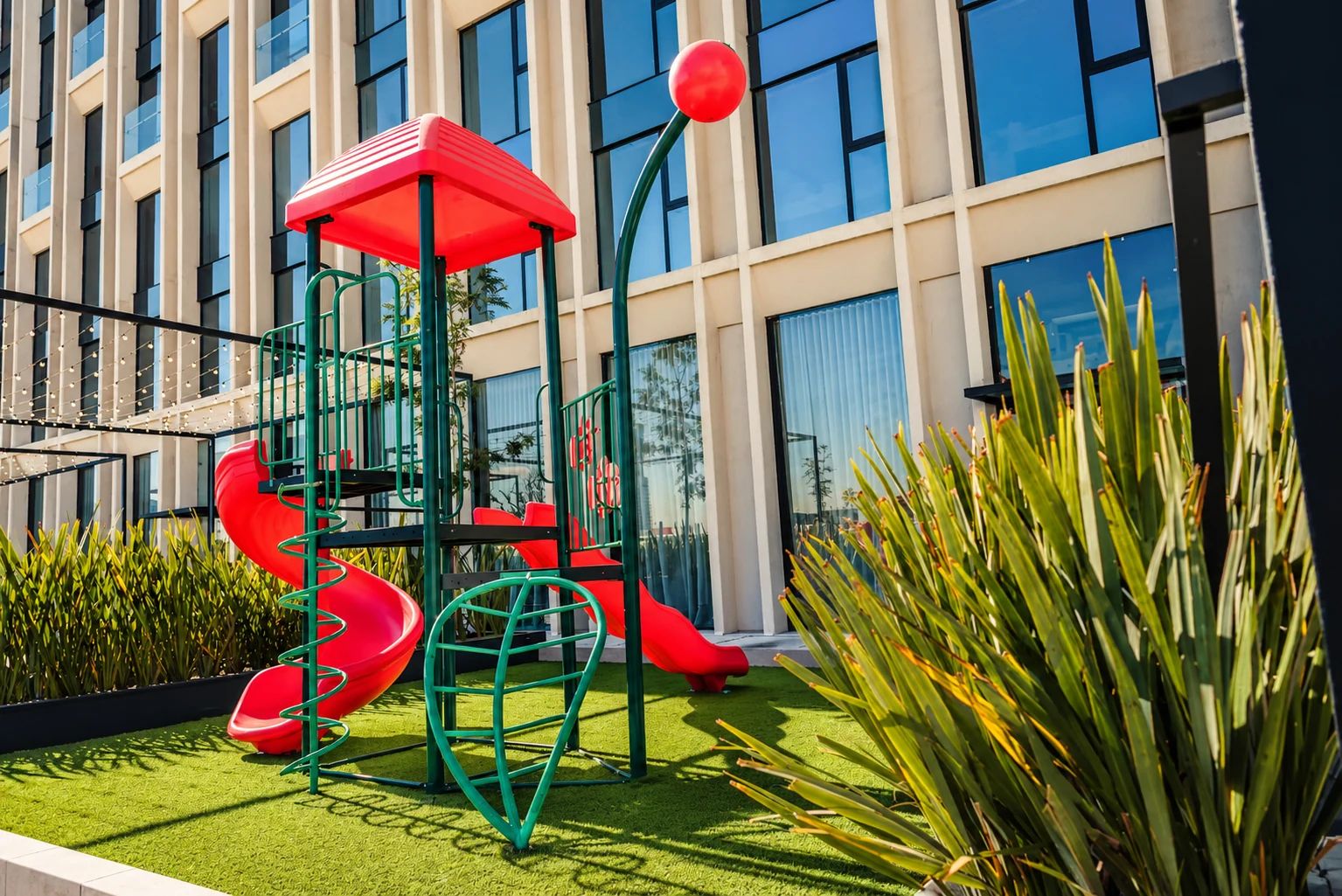A playground with slides and climbing structures, surrounded by greenery and a building with large windows.
