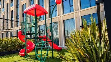 A playground with slides and climbing structures, surrounded by greenery and a building with large windows.