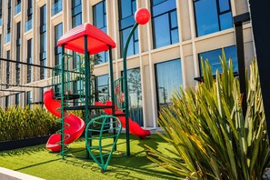 A playground with slides and climbing structures, surrounded by greenery and a building with large windows. - APT21 Puebla - Volcano, Ferris Wheel & City Views in Torres Boudica (San Andrés Cholula,)