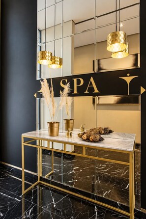 Zona de recepción de spa con una mesa consola dorada, pastos de pampa en macetas doradas y un cartel que dice 'SPA'.