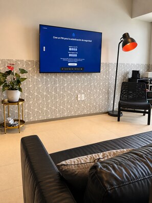 A modern living room with a flat-screen TV mounted on the wall, a black leather sofa, a small side table with a potted plant, a floor lamp, and a patterned wallpaper. - APT21 Puebla - Volcano, Ferris Wheel & City Views in Torres Boudica (San Andrés Cholula,)