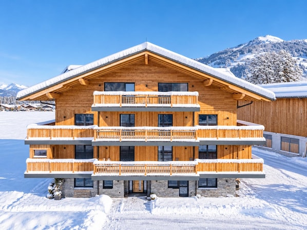 A multi-story wooden chalet with balconies, set against a snowy mountain backdrop.