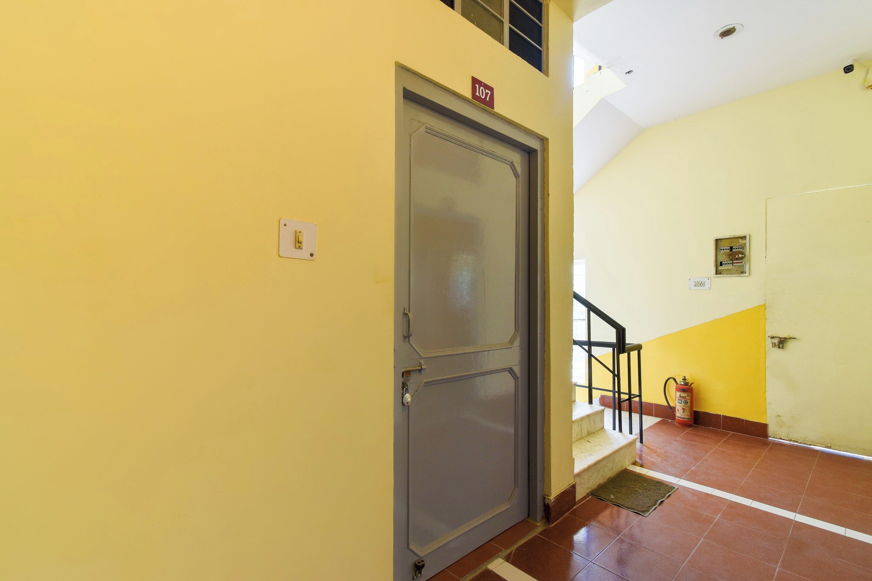 A hallway with a door marked '107', a fire extinguisher, and a staircase with a handrail.