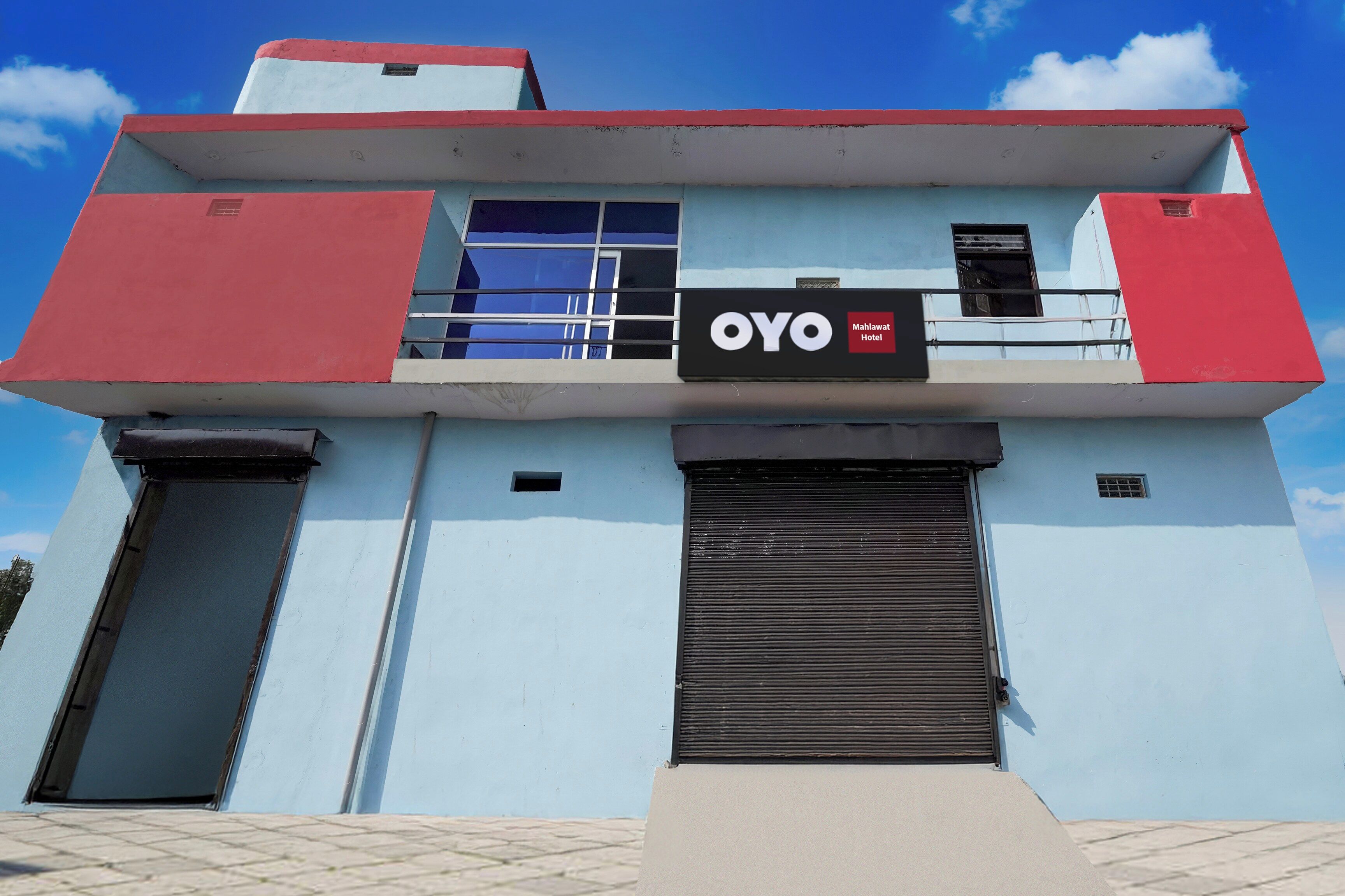 Hotel com fachada vermelha e azul, uma placa preta com os dizeres 'OYO' e 'Mahlawat Hotel', e entrada com persiana fechada.