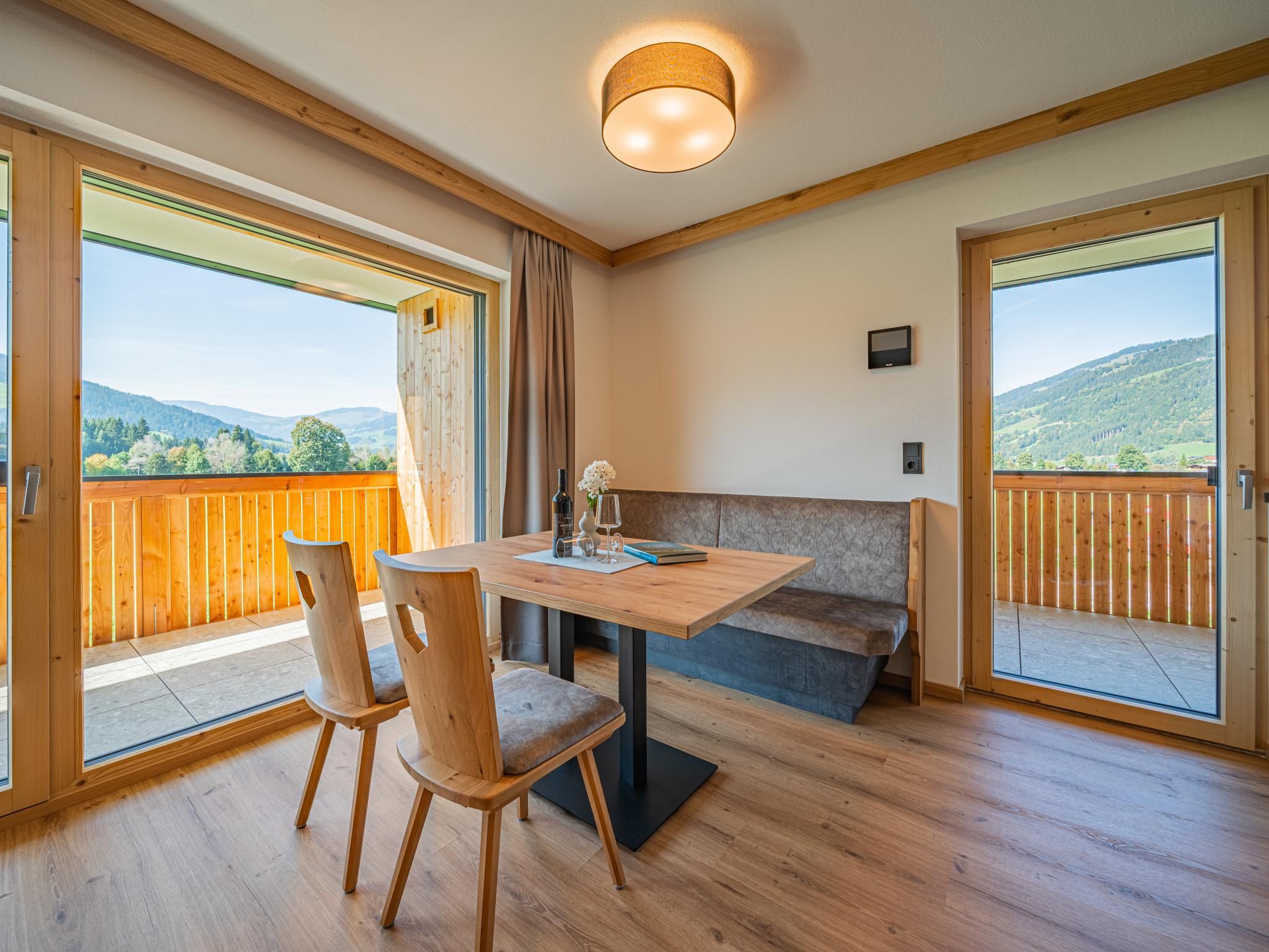 Une pièce avec une table en bois, deux chaises, un banc et de grandes fenêtres donnant sur l’extérieur.