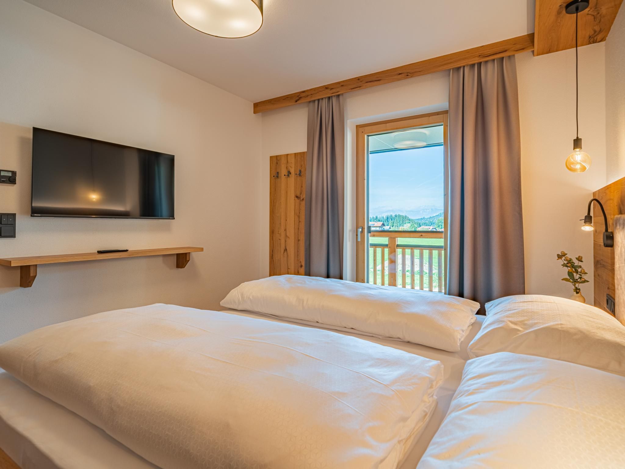 Ein Schlafzimmer mit einem großen Bett, einem Wand-Fernseher und einem Fenster mit Blick auf einen Balkon und viel Grün.