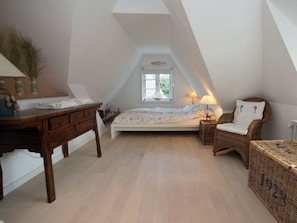 Ein Schlafzimmer mit schrägen Wänden, einem Bett mit floralem Bettzeug, einem Holztisch, einem Korbstuhl und einem kleinen Nachttisch.