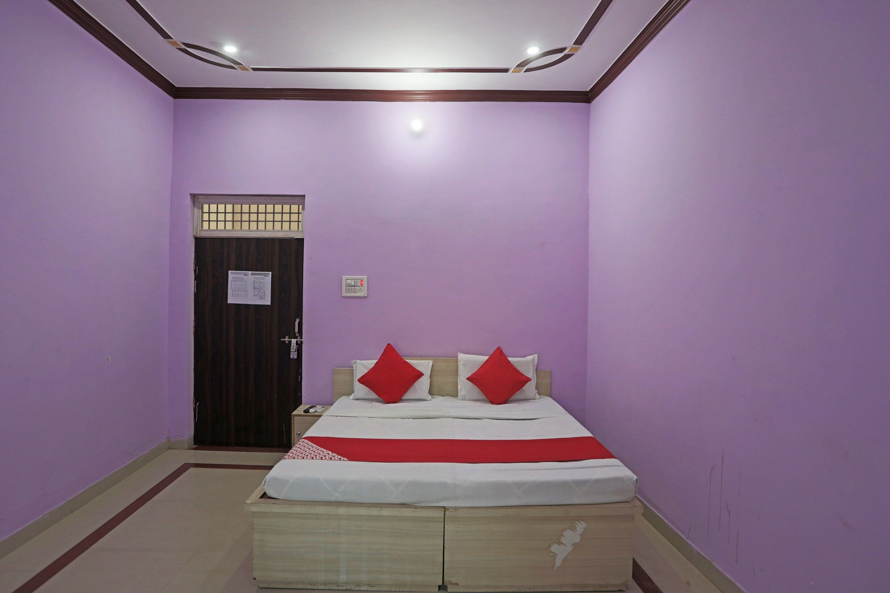 Una habitación con paredes moradas, una puerta de madera, una cama con colcha blanca y roja, y una mesita de noche con un teléfono.