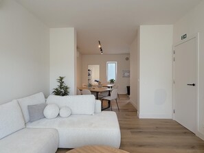 A modern living room with a sofa, a dining area with a table and chairs, and a hallway leading to a door. - Stylish stay near the harbor and beach (Blankenberge)