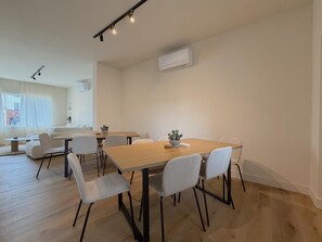Een moderne, goed verlichte eetruimte met een houten tafel en witte stoelen, een kleine plant op de tafel, en een airconditioner aan de muur.